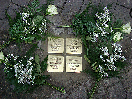 Vier Stolpersteine angeordnet im Quadrat umkränzt von weißen Rosen mit lamngen Stielen