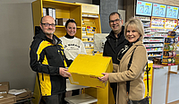 Ein Mitarbeiter in Deutsche Post-Uniform hält gemeinsam mit der Oberbürgermeisterin ein gelbes DHL-Paket als würden sie es nun an die Filialleiterin hinter dem Schalter übergeben. Der Ortsvorsteher steht zur Rechten der Oberbügermeisterin.