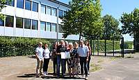 Auf dem asphaltierten Bereich der Grundschule Lauterbach stehen neun Personen, die Oberbürgermeisterin und der LAG Warndt-Saargau Vorsitzende halten den Förderbescheid in Händen